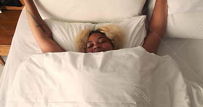 Top view of a young African American woman lying in bed under a white blanket.