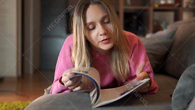 A blonde girl is relaxing at home in the living room. Happy woman reading a magazine lying on a sofa in the living room