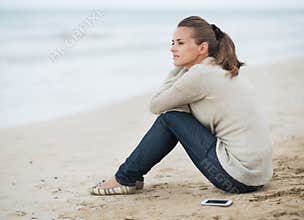 Young woman in sweater with cell phone sitting on lonely beach