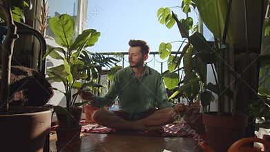 Young hipster guy takes care of flowers. A man sits down in center of room in home. The concept of plant care and
