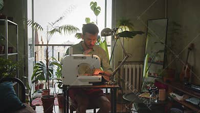 Male clothes designer works at sewing machine from home. Young entrepreneur starts sewing business at home in room with