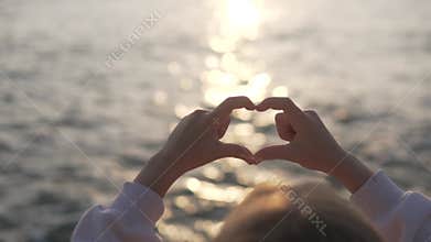 Little girl showing with her fingers a heart symbol on the sea with sunset