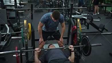 Top view of personal fitness trainer helping beginner sportsman doing barbell bench press exercise during workout in gym