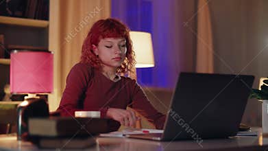 In her cozy room, a young woman with red hair is deeply focused on studying with her laptop