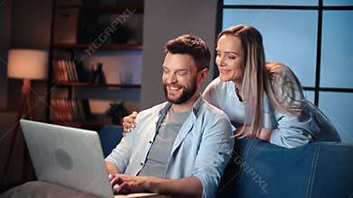 Happy spouses at home in evening, looking at laptop display, viewing pictures in social media online