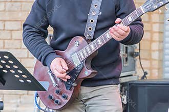 A person is playing an electric guitar, seated and focused, wearing a dark top and light pants Microphone stand suggests