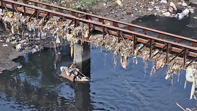 5-7-24 A polluted canal, surrounded by garbage, with a bridge pillar above.
