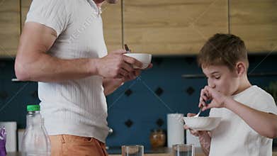A friendly family of father and son holds bowls in hands and eats, standing in the kitchen. A dad shares an idea for a