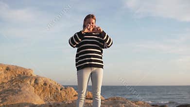 A woman shows her heart with her hands while standing on a rock by the sea