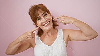 Cheerful middle age woman points to perfect teeth, flaunting dental health over a pink isolated background