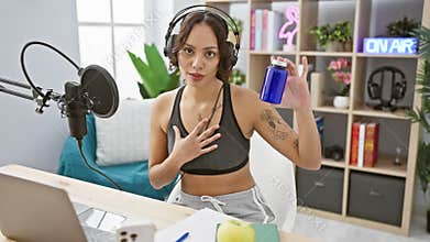 Young woman podcasting indoors with microphone and headset in a modern studio setting, holding a blue bottle