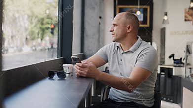 A bald man is sitting at a cafe window, using his smartphone with a contemplative expression
