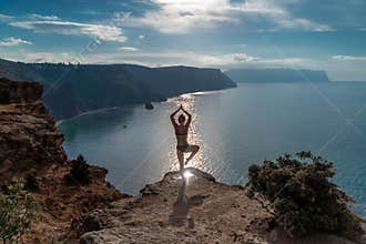 A woman is doing yoga on a cliff overlooking the ocean. The scene is serene and peaceful, with the woman& x27;s pose and