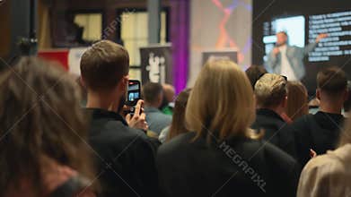 A man with beard and blonde girl are sitting at lecture among a large group of people. In workshop brimming with