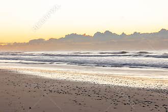 Sunrise behind a cloudbank in a long exposure photo, reflects its glow on the shallow water.