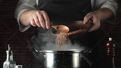 A chef in a black apron spoons buckwheat from a bowl into a hot pot, while kitchen utensils and a brick wall create a warm