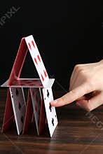 Woman destroying house of playing cards at wooden table against black background, closeup