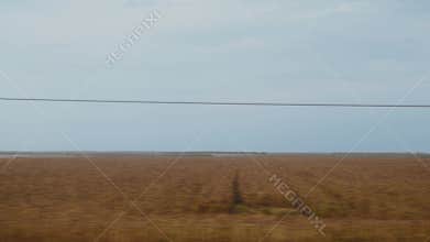 A vast, flat countryside landscape from a moving train. A dirt road cuts through harvested fields under a pale sky. Expansive and