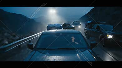 Nighttime convoy of vehicles driving on a winding mountain road with headlights illuminating the path