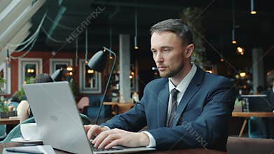Serious young man in suit using laptop typing working alone in restaurant
