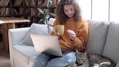 Relaxed hispanic teen girl using laptop watching movie playing with cat on sofa.