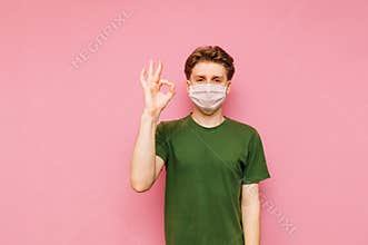 Positive guy in a medical mask isolated on a pink background, looks at the camera with a smile and shows a OK gesture. Cheerful