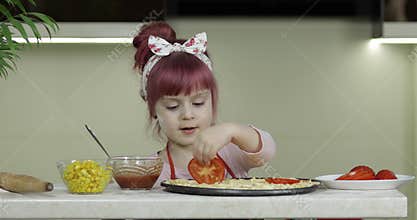 Cooking pizza. Little child in apron adding sliced tomatoes to dough in kitchen