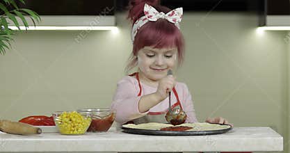 Cooking pizza. Little child in apron adding tomato sauce to dough in kitchen