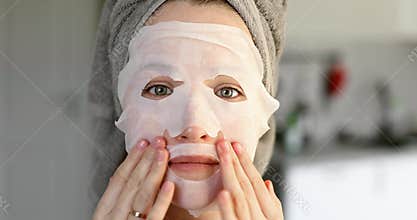 Young girl with towel on hairs applying cosmetic sheet mask on face, front view.