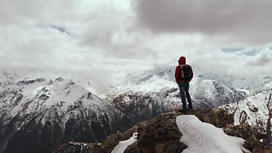 A male traveller with a backpack stands on top of a cliff against the backdrop of snow-capped mountain peaks and low