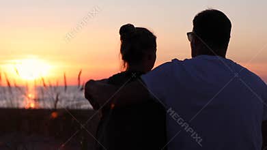 young lovers couple sitting in sand on beach at romantic golden sunset. back view
