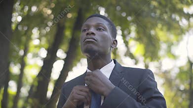 Close-up of young handsome African American man adjusting necktie in summer park and leaving. Portrait of confident