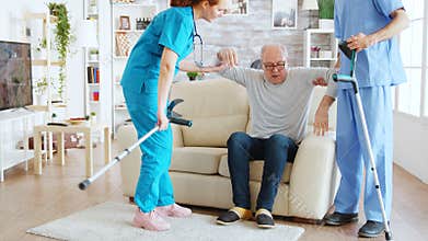 Team of nurses or social workers helping an old disabled man to walk with his crutches out of the nursing home room