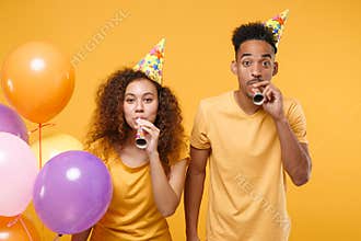 Cheerful young friends couple african american guy girl in birthday hat isolated on yellow background. Holiday party