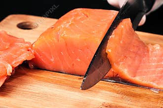 Hands cooks close-up. The chef cuts with a knife a red fish, smoked salmon on a wooden cutting board.