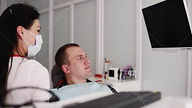 Caucasian female dentist in white robe, mask and black gloves explaining x-ray image on the black screen to male patient