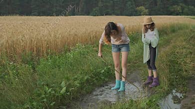 Two happy girls sisters in boots playing in puddle of rainwater on country road