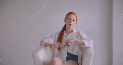 Closeup portrait of young pretty caucasian redhead female looking at camera holding her glasses with thoughtfulness