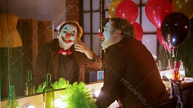 A crazy man clown in the dressing room applying his paint - drawing a smile with a red paint and smile