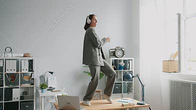 Beautiful office worker in suit dancing on desk in office wearing headphones