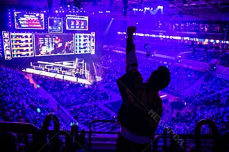 Big esports event. Video games fan on a tribune at tournament`s arena with hands raised. Cheering for his favorite team.