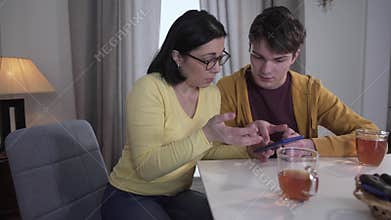 Teen brunette Caucasian boy teaching adult mother in eyeglasses to use smartphone. Patient son helping woman to deal