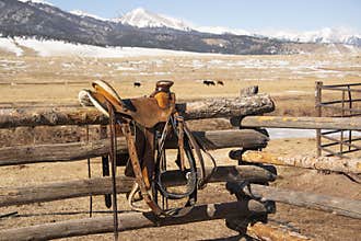 Western leather riding saddle with stirrups and pommel horn on rustic wood ranch fence