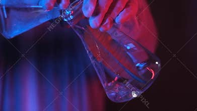 Scientist in neon blue light pours a clear solution into a beaker.