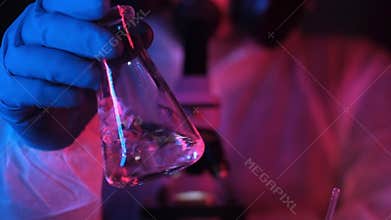 Scientist or chemist in a laboratory in neon light shakes liquid in a beaker.