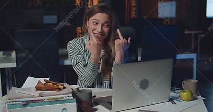 Young woman having video call with boss on her laptop in night office. Angry female employee showing fuck you after