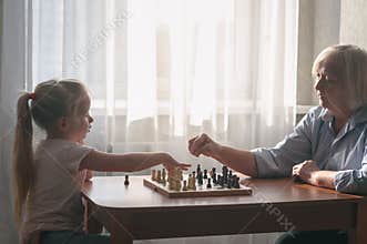 Senior teaches a child to play chess. People are sitting at a table by the window. On the table a chessboard with pieces.