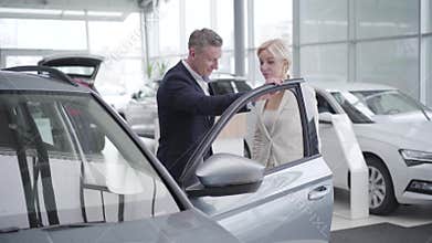 Car dealer opening car doors for successful adult Caucasian businesswoman in showroom. Wealthy female client choosing