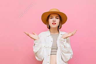 Funny woman in a hat looks puzzled at the camera with raised arms from the bewildered face on a pink background. Portrait of