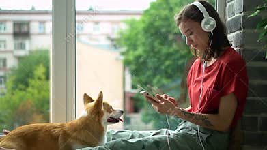 Hipster millennial girl listen to music in headphones and stroke corgi dog Avki. happy 20s smiling